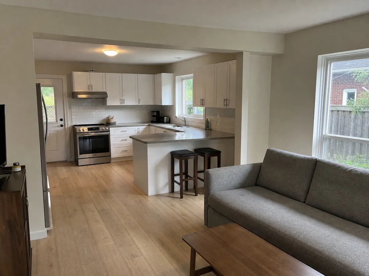 A kitchen opened up to the living room through a wide new opening with a flush beam, white cabinets on one side and a grey sofa on the other